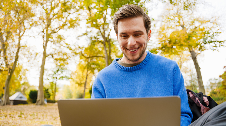 Smiling man in bright blue sweater sits outside on his computer